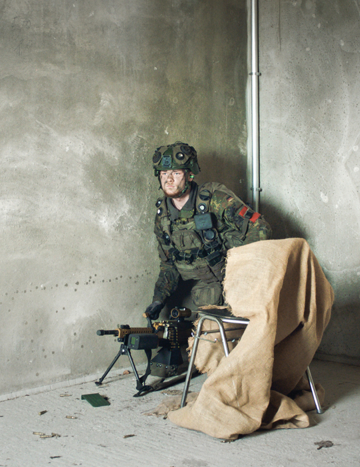 A soldier in uniform crouches inside an empty concrete room with a machine gun