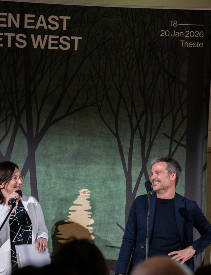 A light-skinned woman with dark hair and glasses, wearing a black shirt and a white cardigan talks into a microphone while talking to a white man in a black sweater with short-cropped hair with a poster advertising When East Meets West