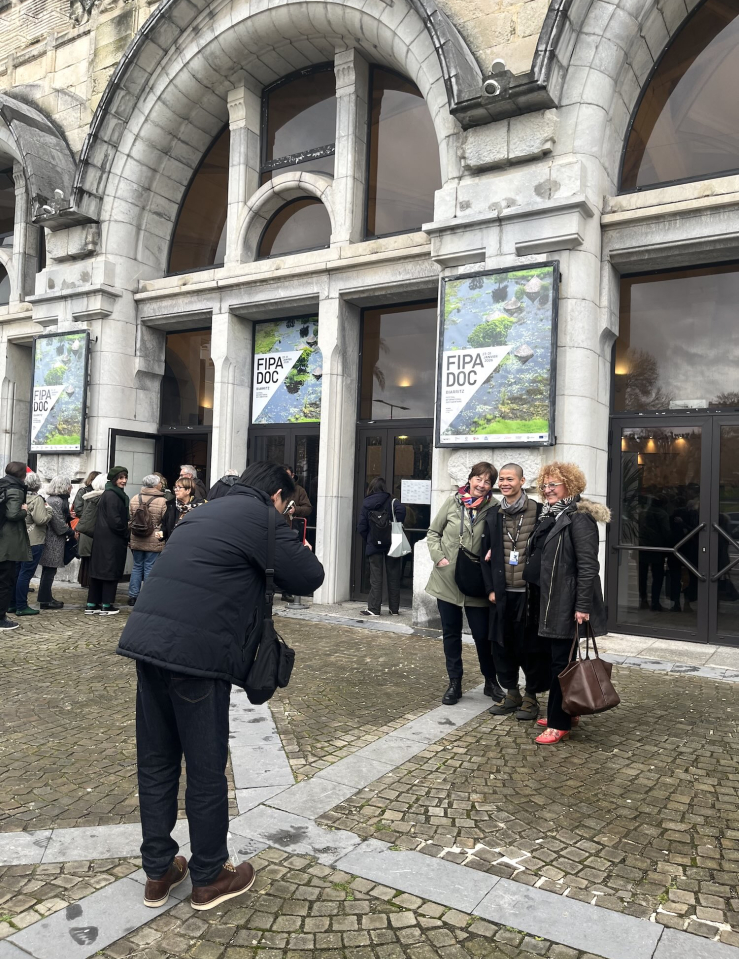 Outside of a theater, a man in black clothes takes a photo of another man posing with two older white women.
