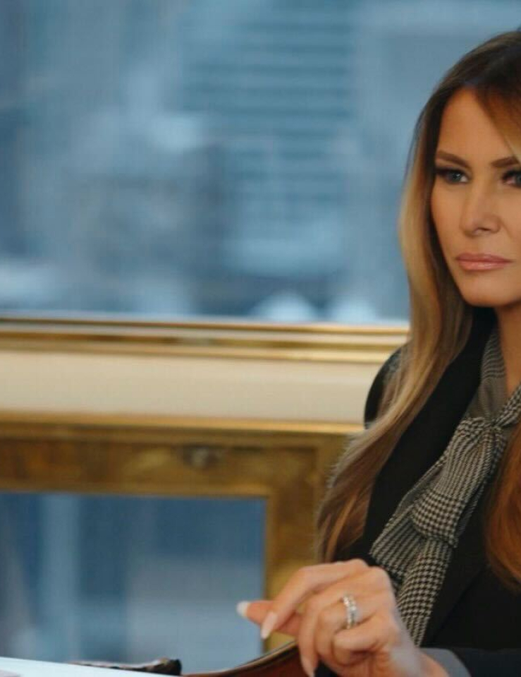 A heavily-made up white woman with long, colored blond hair sits a desk with a stern look on her face