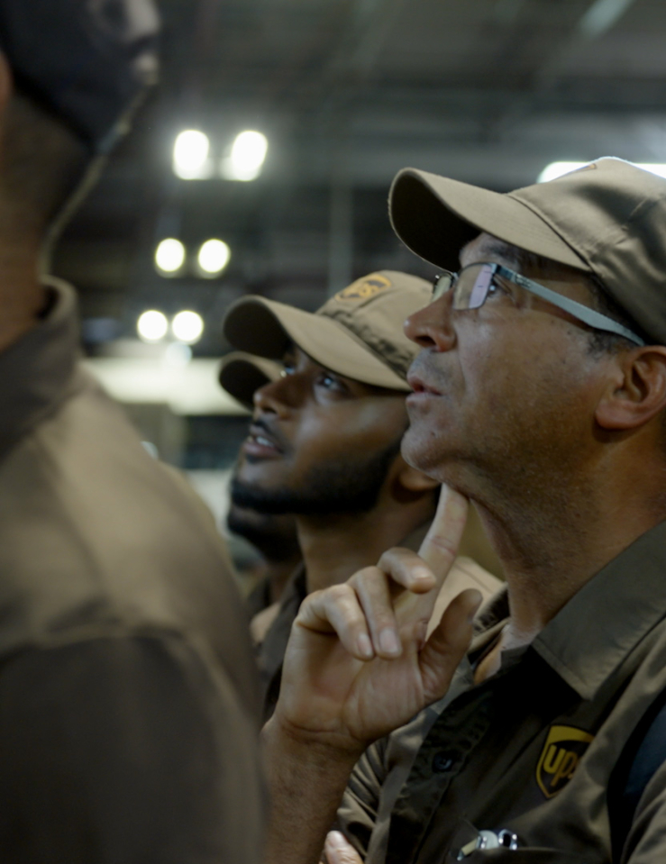 A group of men dressed in brown UPS uniforms look up, while standing inside a warehouse.