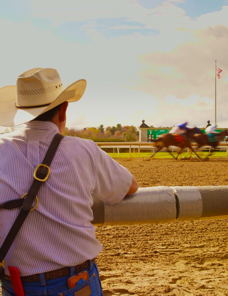 A man in a cowbow hat seen from behind is watching a horse race happening across from him