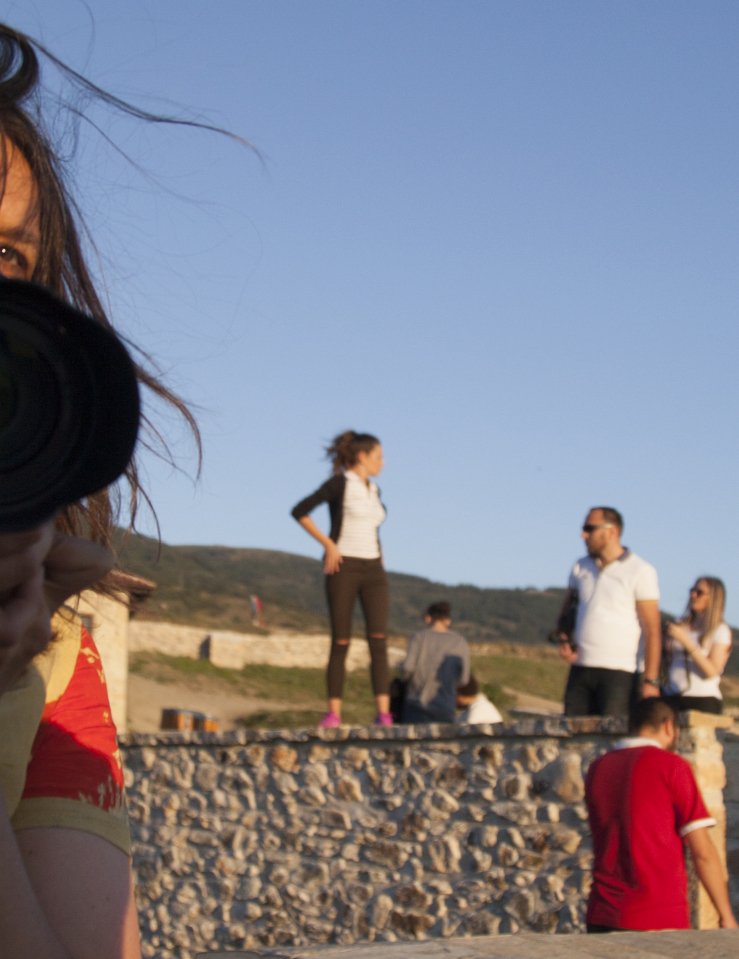 A white woman holding a camera is seen in a mirror as a group of people stand along in the background in a desert field