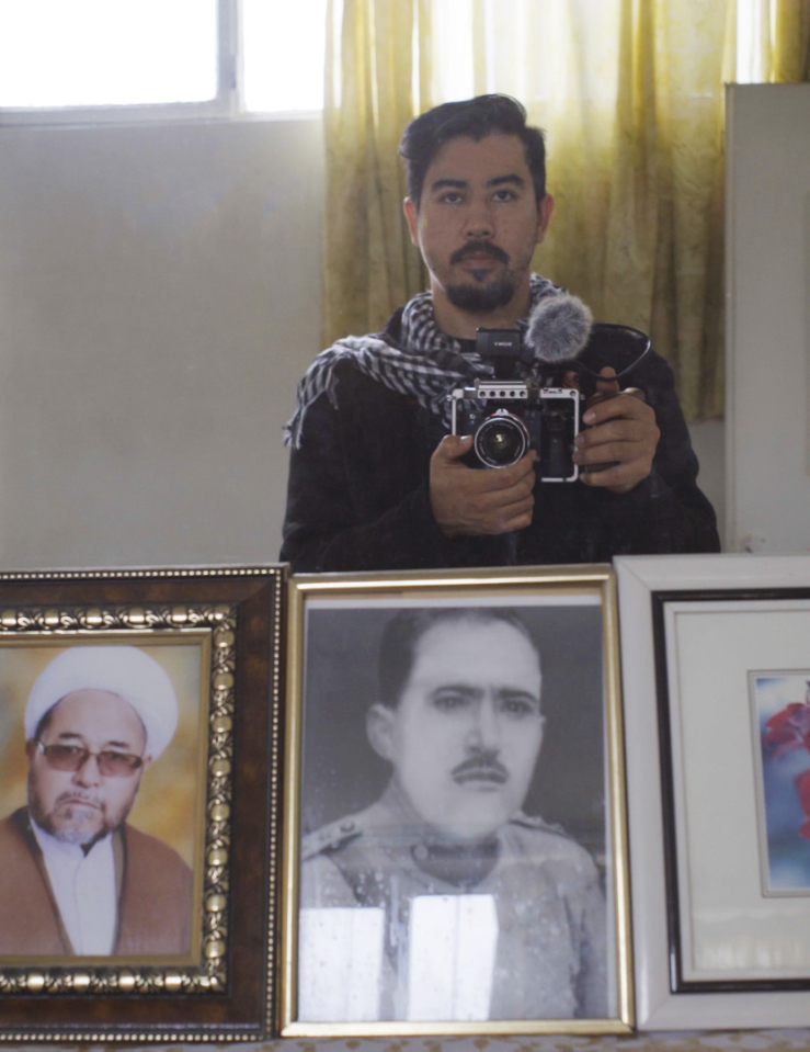 A young brown-skinned man with short black hair and goatee beard holds a camera in front of a mirror where we can see various photos of older Middle-Eastern men in frames.