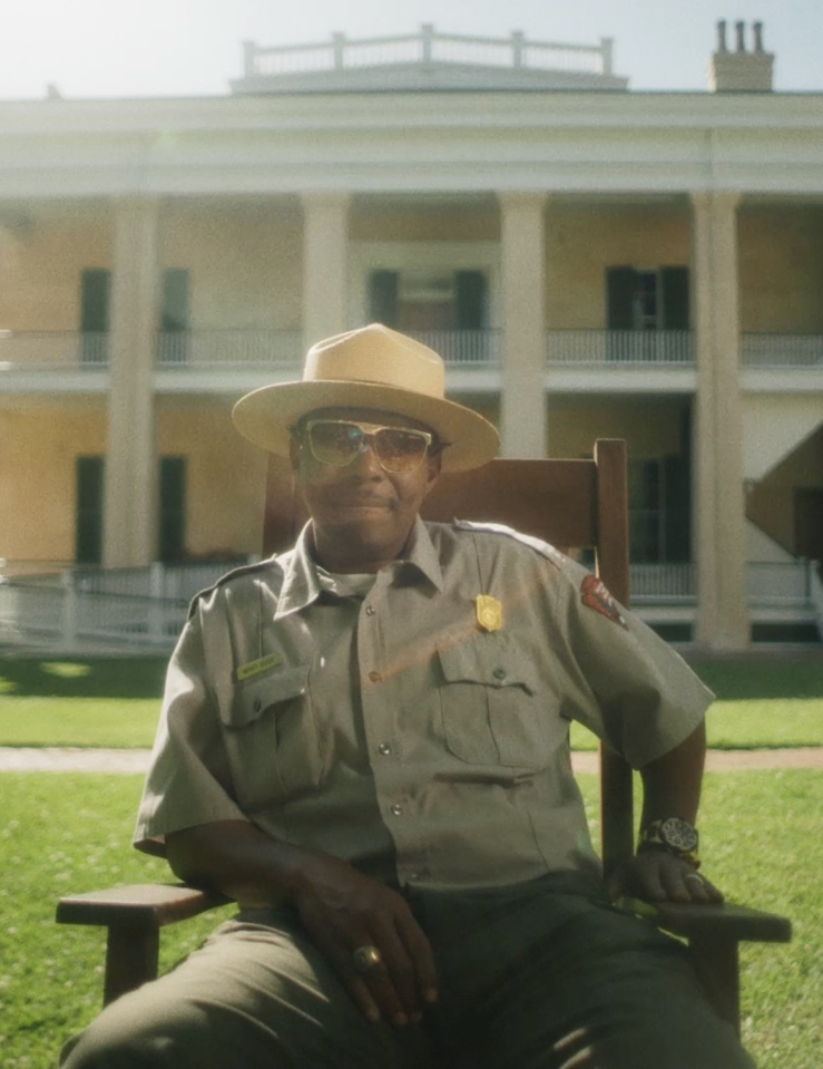 In front of a yellow plantation home, a National Park Service ranger leans causally on one elbow while sitting on a wooden chair.