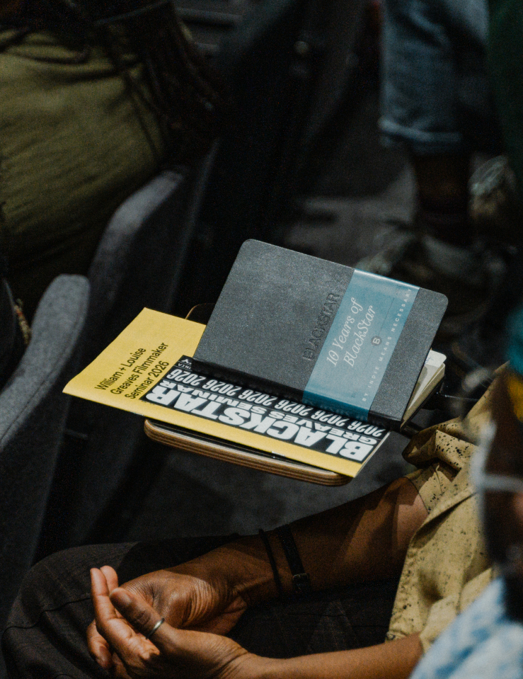 Close up shot of a Blackstar program and notebook on top of a desk, amid a number of Black folks seated as if for a lecture