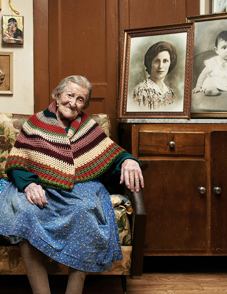 An elderly woman with pulled back grey hair sits on a yellow-print heavy coach surrounded by framed black and white photos of her younger self 