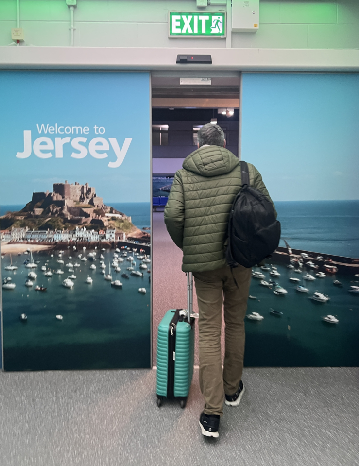 Middle-aged man in warm winter jacket and a backpack with a roller bag heads through some doors that read "Welcome to Jersey"