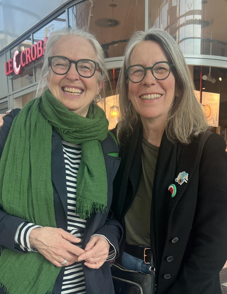 Two middle-aged white women with white auburn hair and rimmed glasses smile at the camera
