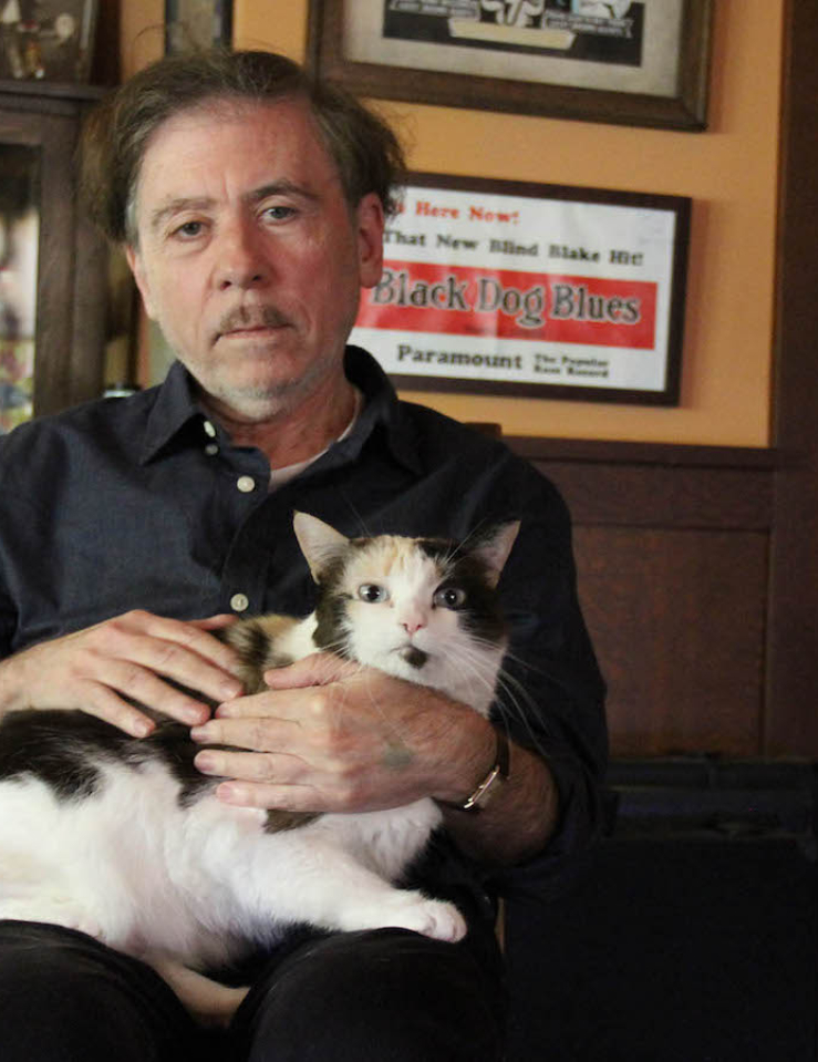 An older white man with thinning black hair and a bit of stubble, wears a dark blue shirt and dark pants as he sits staring at the camera as a white and black cat sits on his lap