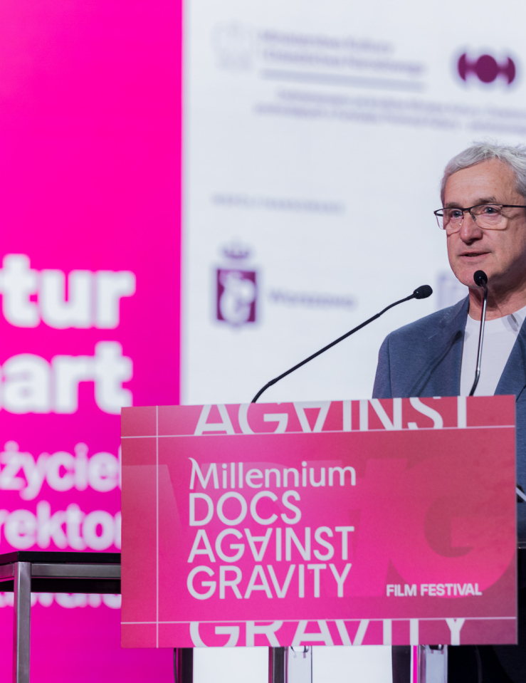 A middle-aged white man with grey hair and glasses wears a suit and a tee while standing at a podium that's branded as Millennium Docs Against Gravity in bright pink