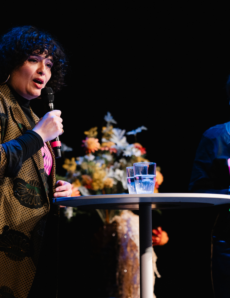 A light-skinned woman with shoulder-length curly black hair in a stylish printed coat holds a mic at a table with two glasses as a young blond-haired woman wearing a CPH:DOX tee stands in the background