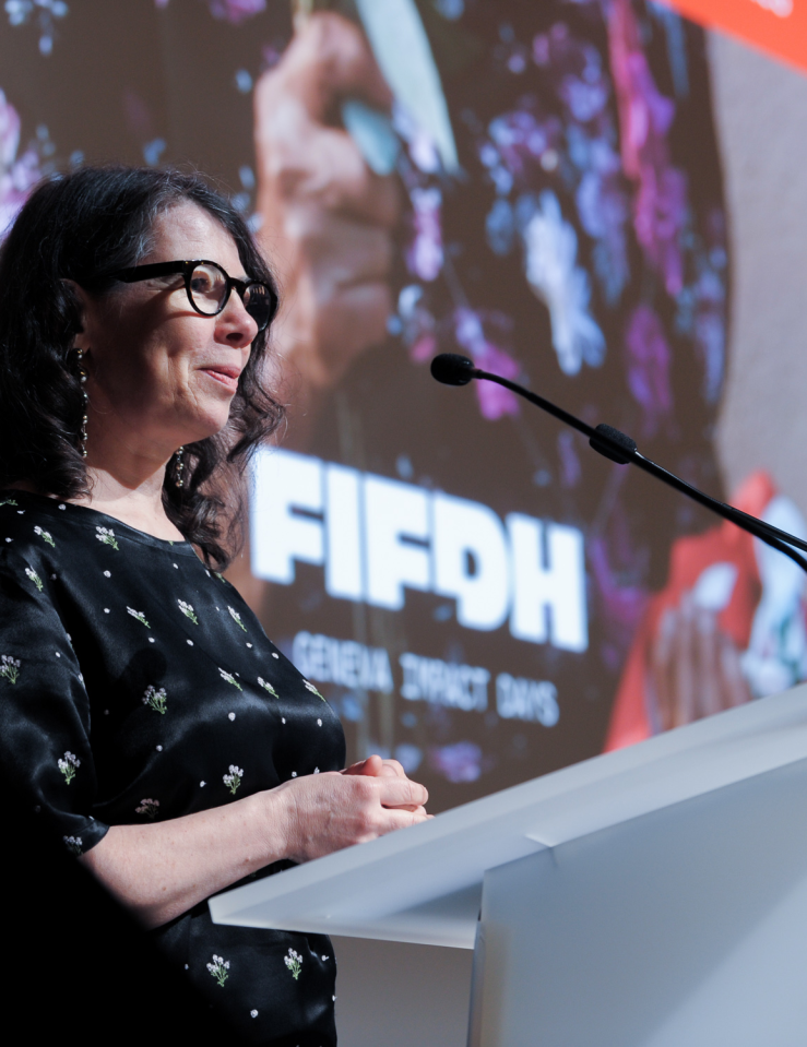 A middle-aged white woman with shoulder-length black wavy hair dons glasses and a black dress with white flowers at a podium with a mic