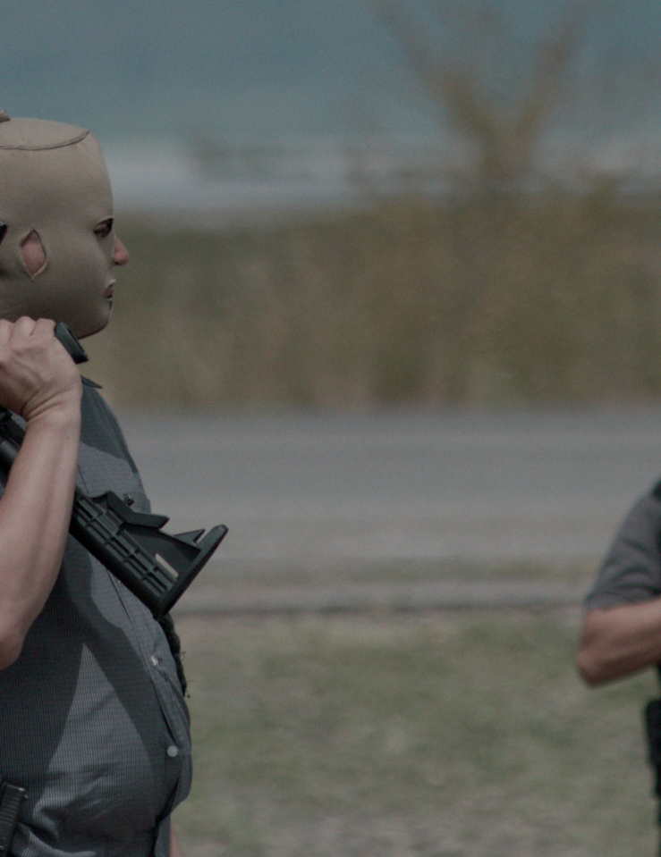 Two men in cammo gear stand in an empty field holding guns don skin-colored masks that reveal only their eyes, nose, and mouth