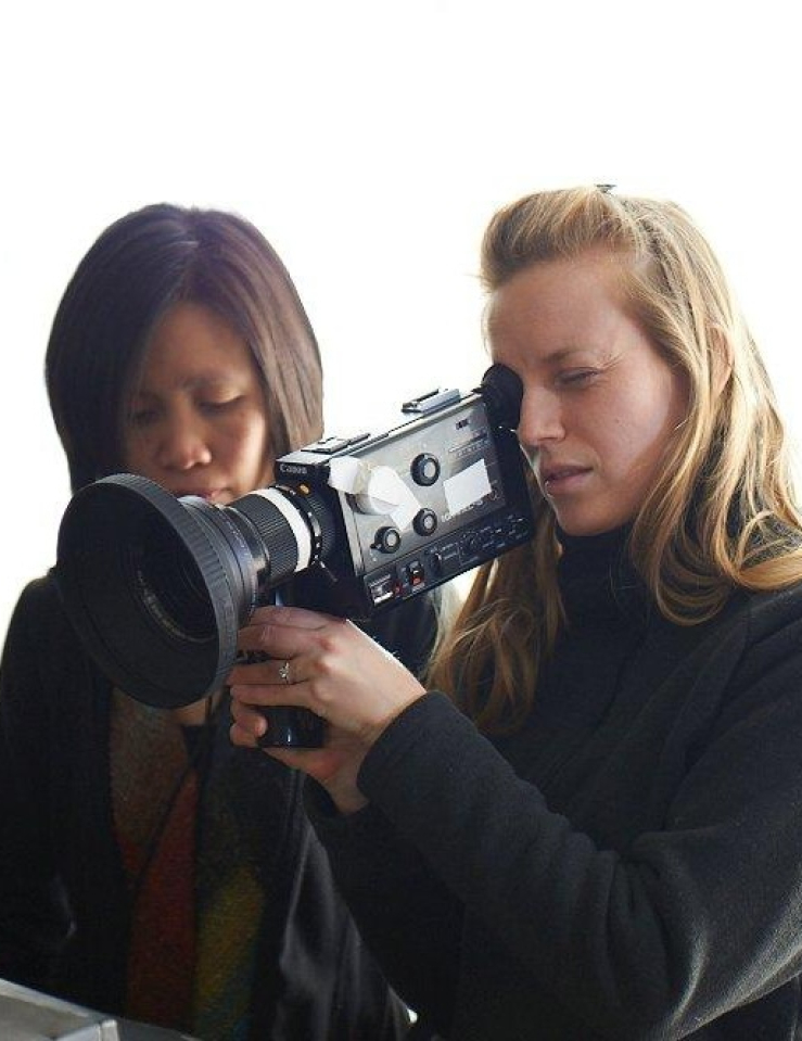 A young blond female filmmaker holds a camera against a bright window behind her; a young Asian woman with dark hair stands behind her
