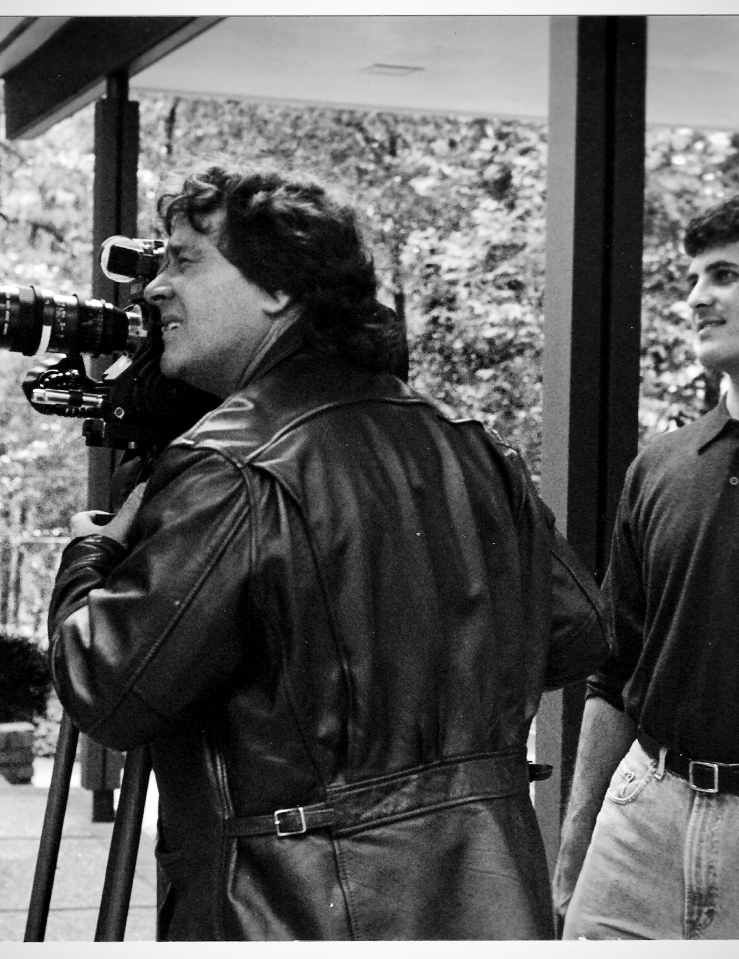 On the left, a man in a black leather jacket peers into a video camera. On the right, a younger man in a tucked polo observes.  Black and white photo