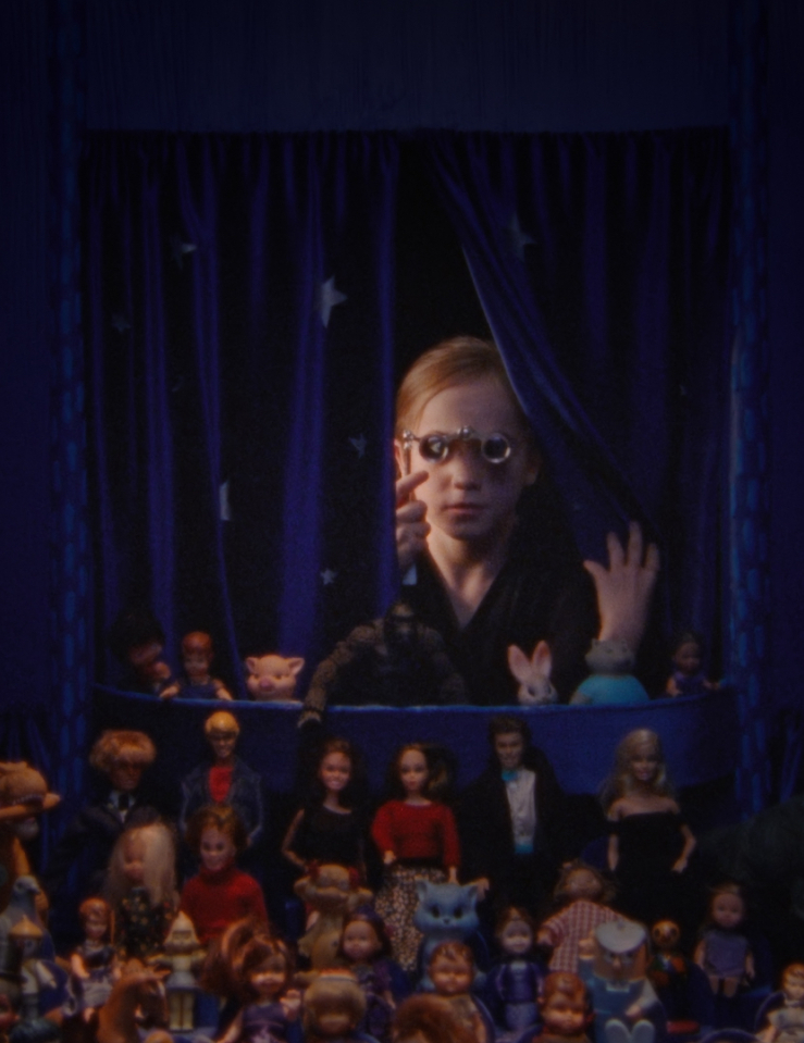 A blonde girl peers through spectacles into a space enclosed in purple curtains filled with various dolls