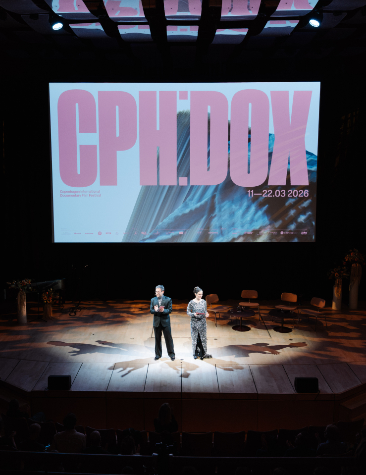 A man in a suit and a woman in a sequined gown stand in the center of a stage, illluminated by a spotlight. A large screen appears behind them which reads "CPH:DOX" in pink text