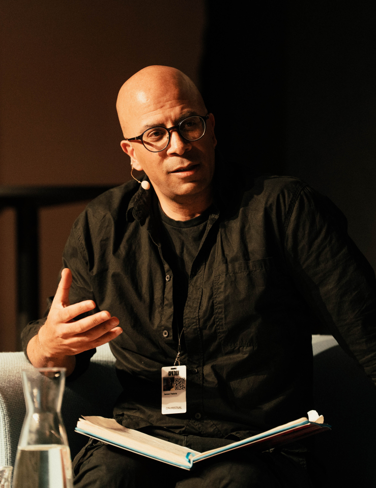 Bald man in glasses and dark brown collared shirt sits in white arm chair, gesturing with hand