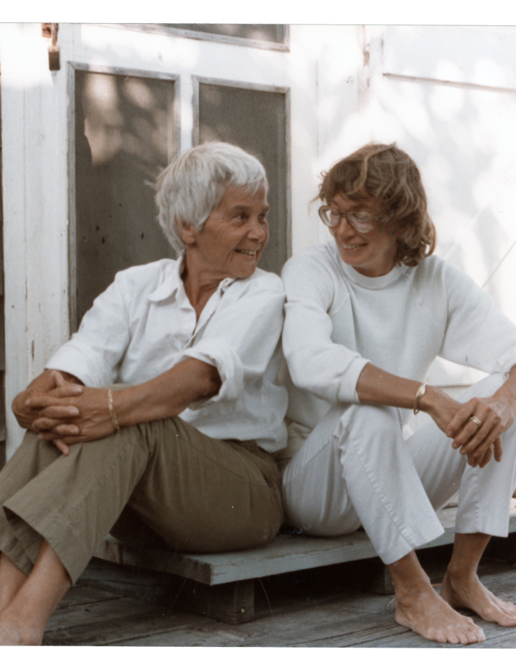 Two women sit on a doorstep smiling at each other. On the left is an older woman with short white hair in khaki pants and a white collared shirt. On the right is a woman with mid-length brown hair, glasses, a white turtleneck and white pants. Both are barefoot