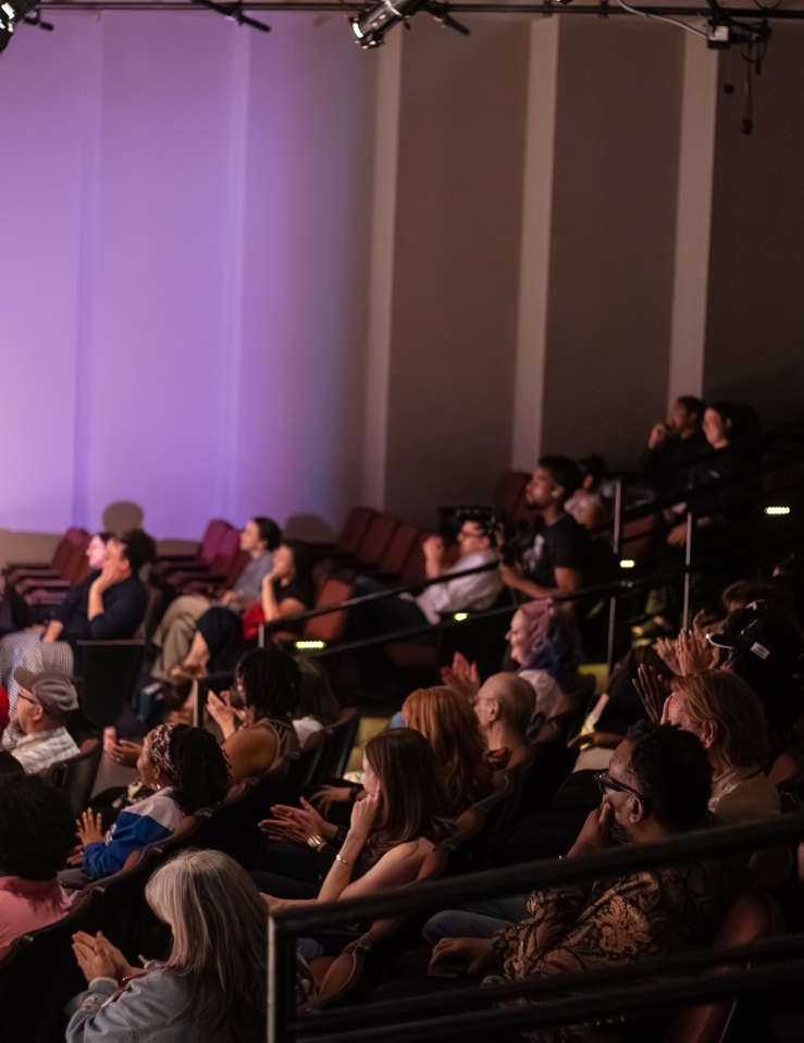 A packed theatre focuses on a stage which is off camera to the left. Purple light illuminates the audience