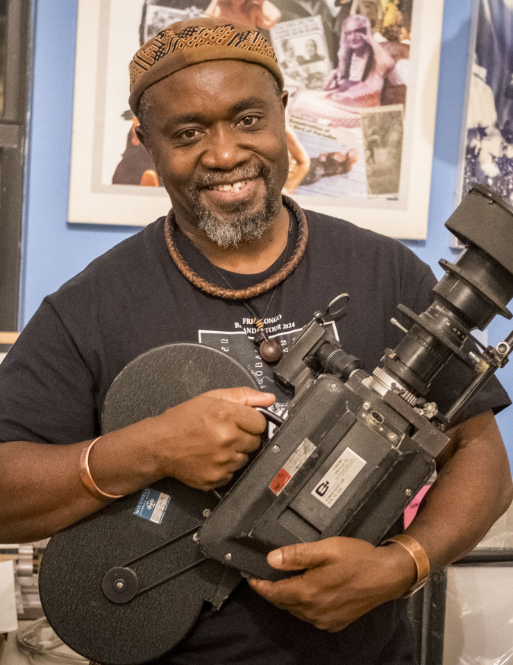 A smiling Black man in a brown hat, black T-shirt, and wooden and metal jewelry holds an old camera in front of awards and posters, one depicting Malcolm X