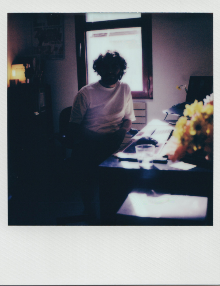 Polaroid of a man in a white T-shirt sitting at a desk which houses yellow flowers. The man is backlit by a window behind him and his face is obscured