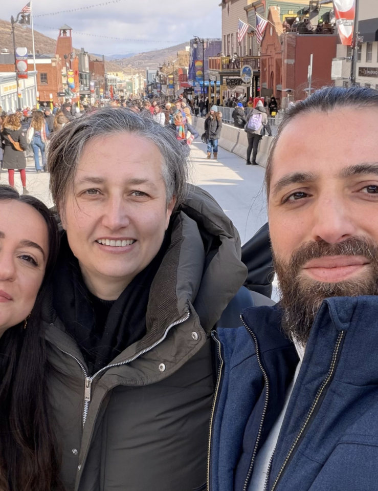 Selfie taken by a bearded man (right), woman with short grey hair (center), and woman with long, dark hair (left). All wear warm coats and smile. The background is a street lined with shops and pedestrians