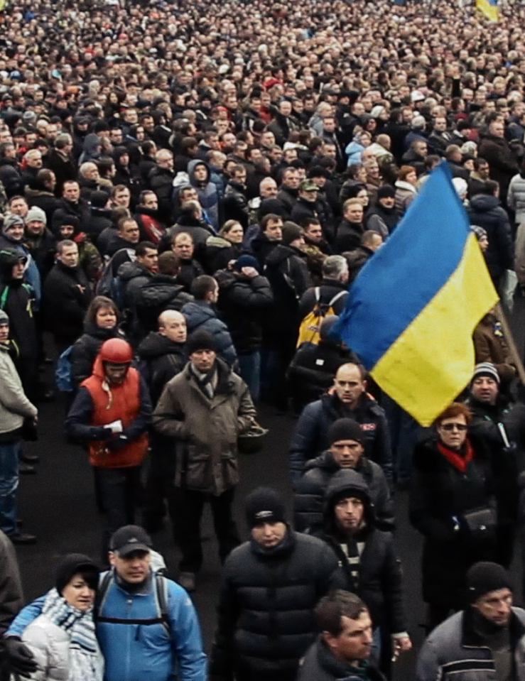 A massive crowd of people in winter clothes is gathered. One man towards the front waves a large Ukrainian flag