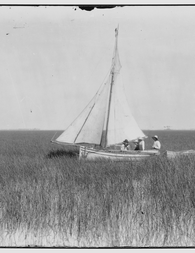 Black and white image of three men sitting in a white sailboat amidst marshland