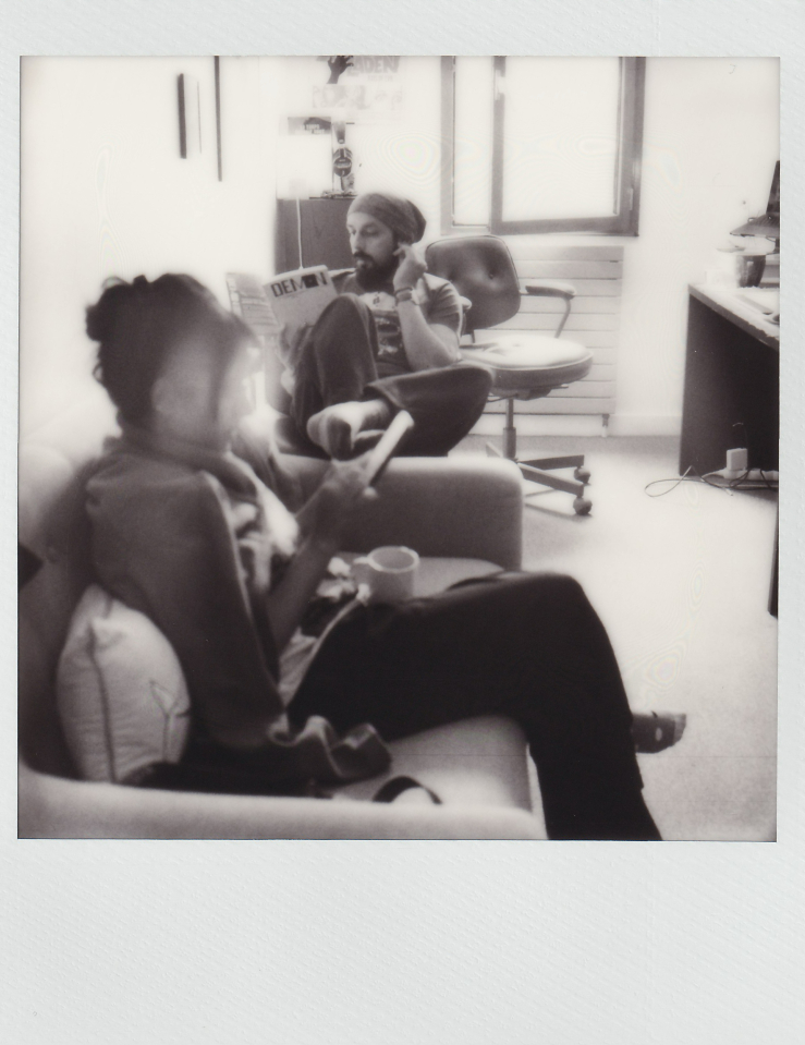 Black and white polaroid of a man and a woman sitting in armchairs in a small room. The man reads a magazine and the woman looks at a tablet and holds a mug. A desk and open window can be seen in the background