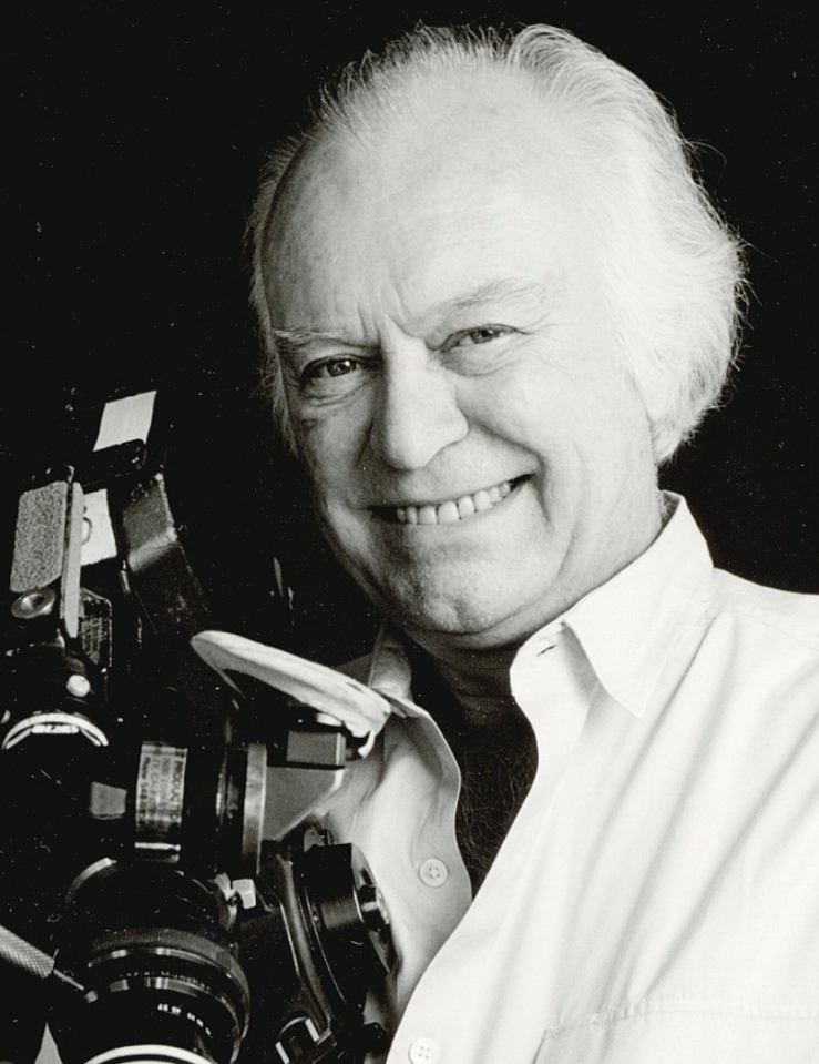 Black and white headshot of an older white balding man with tufts of white hair who smiles at the camera while holding a film camera
