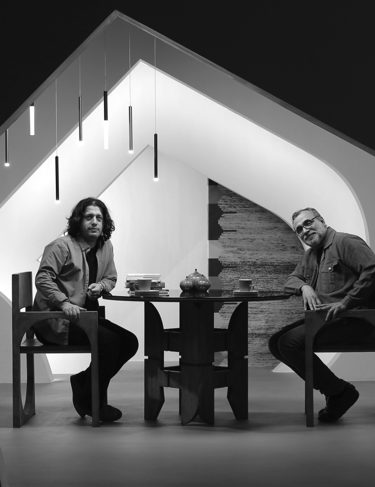 Two men sit at a table in a modern room framed by an unusually shaped doorway. Both look towards the camera. The man on the left has shoulder length dark hair and the man on the right has short greying hair and facial hair and glasses. Two mugs of coffee, a stack of books, and three decorative vases sit on the table. An abstract chandelier appears above the men's heads. Black and white image