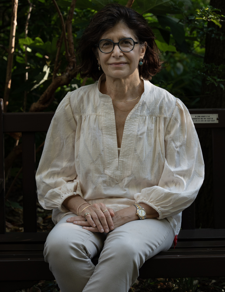 Woman with shoulder length dark hair in glasses and a white blouse sits smiling on a bench in front of a tree