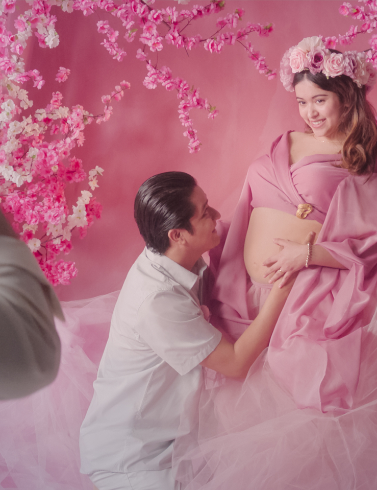 A photographer in frame right photographs a pregnant woman in a pink dress and pink flower crown in front of a pink background. A man in white kneels and kisses her stomach