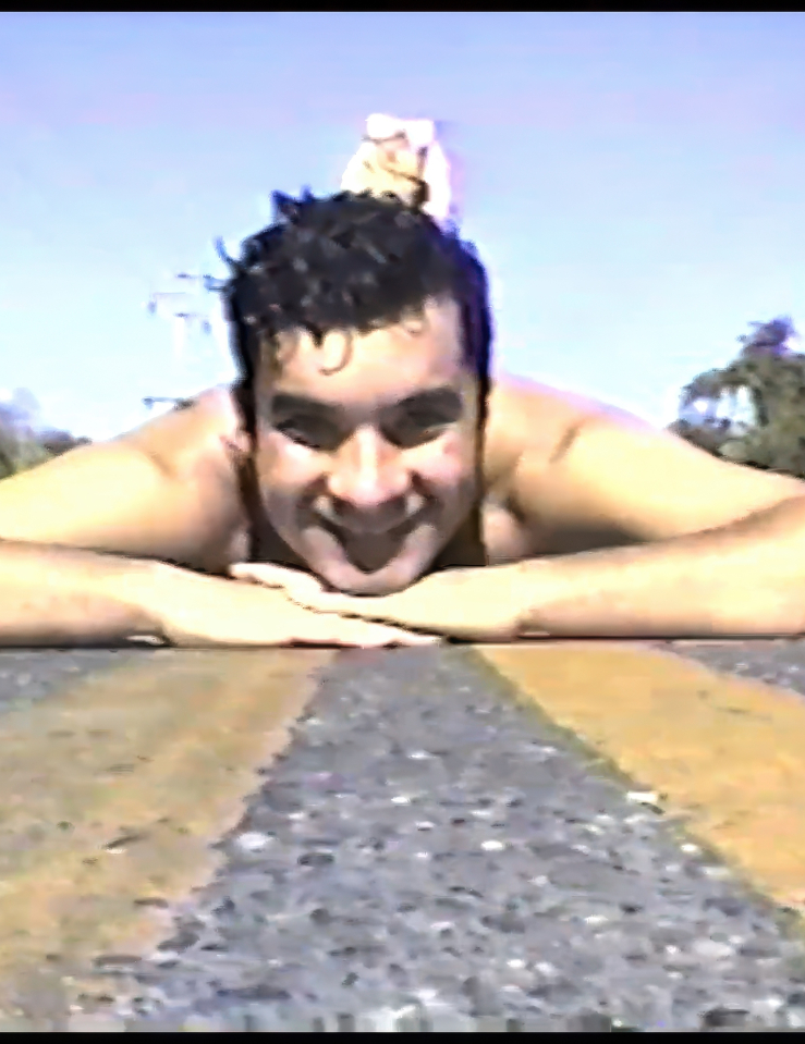 Grainy, digital photo of a smiling, shirtless person lying on the yellow painted lines of an asphalt road. Their chin rests on their hands and their feet appear to be raised behind their head