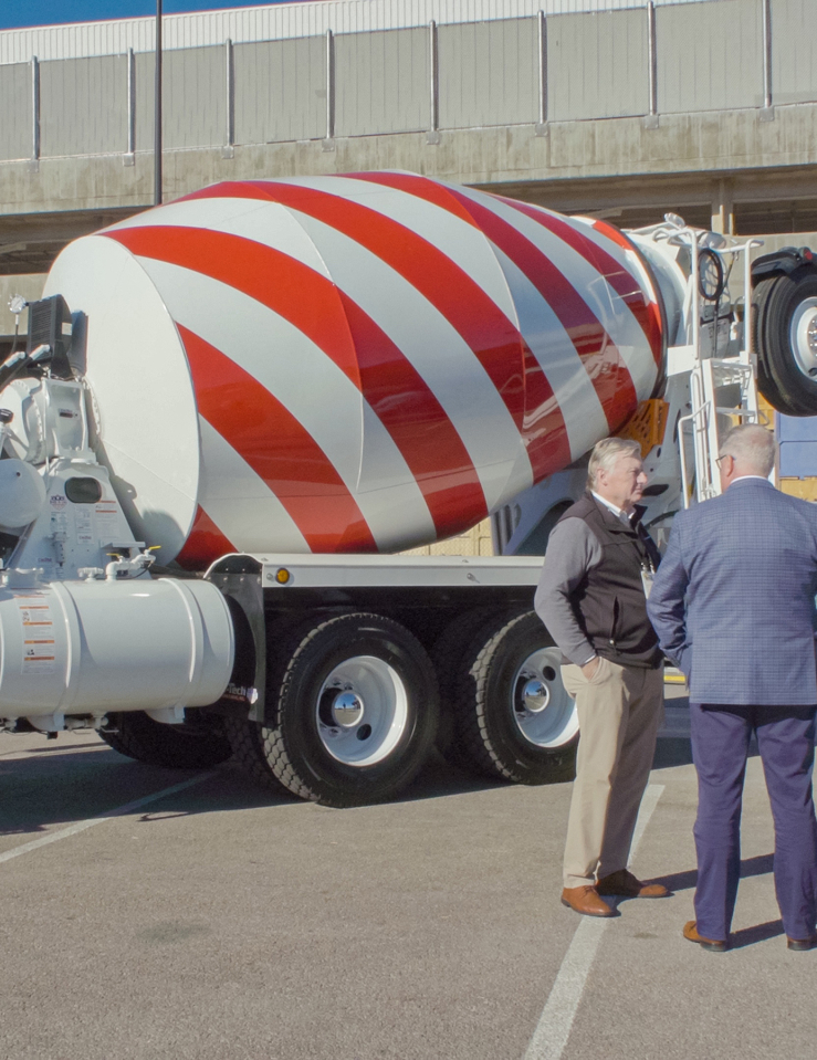 A concrete truck is seen parked on a concrete parking lot, with two white men, one in a suit, standing right by it