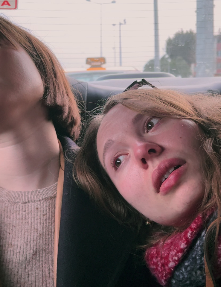 Two women in the backseat of a car, each with mid-length brown hair. The woman on the left, wearing a brown sweater under a black overcoat, sleeps, and the woman on the right, wearing a red scarf, rests her head on the other woman's shoulder and looks out the window